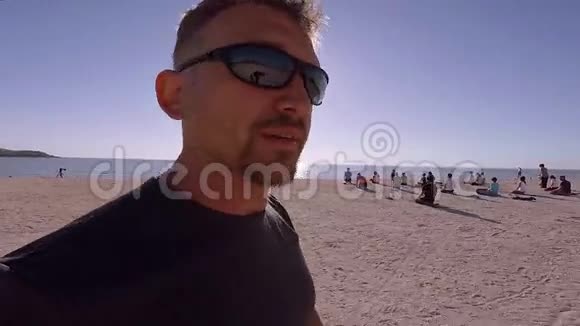 高加索人对着镜头说话在一群从事瑜伽的人中间沿着海滩散步视频的预览图