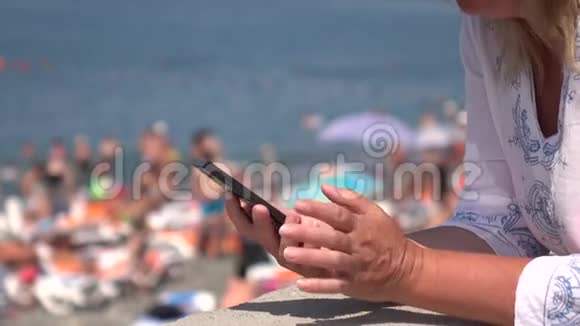 年轻的女人坐在海滩上使用智能手机视频的预览图