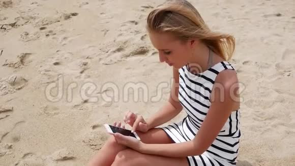 年轻漂亮的女士身材苗条一头金发穿着黑白连衣裙坐在海滩上使用智能手机视频的预览图