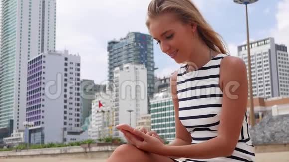 年轻漂亮的女士身材苗条一头金发穿着黑白连衣裙坐在海滩上使用智能手机视频的预览图