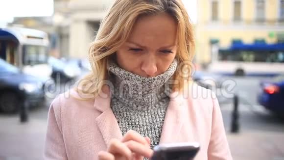 一个穿着粉红色外套和毛衣的疲惫的女人站在拥挤的街道中间看着她的智能手机视频的预览图