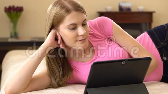 美丽迷人的年轻女子躺在沙发上在平板电脑上看电影笑着视频的预览图