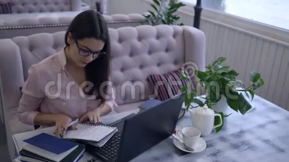 远程学习女学生用钢笔在笔记本上写笔记坐在书桌上笔记本电脑在室内视频的预览图