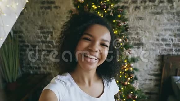 年轻的非洲裔美国女孩在家中圣诞树附近使用智能手机摄像头在线聊天视频的预览图