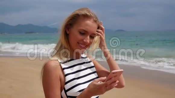年轻漂亮的苗条女人一头金发穿着黑白连衣裙站在海岸上用智能手机视频的预览图
