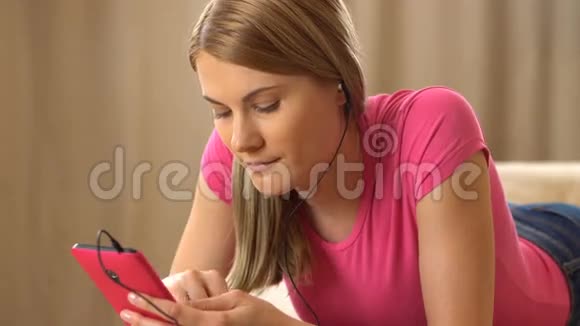 美丽迷人的年轻女人穿着一件粉红色的t恤一部粉红色的智能手机躺在沙发上听着音乐视频的预览图