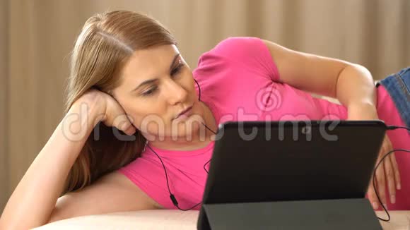 美丽迷人的年轻女子穿着一件粉红色的t恤躺在沙发上用她的平板电脑和视频的预览图