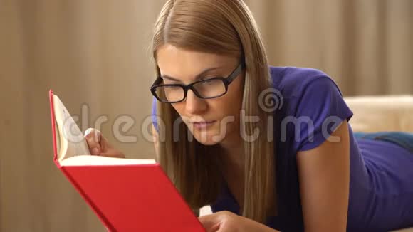 美丽迷人的年轻女人穿着紫罗兰色的t恤和一副黑色眼镜躺在沙发上看书希望视频的预览图