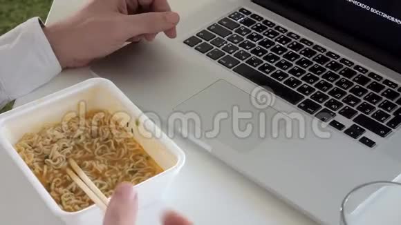 一个男人坐在桌子旁吃中国面条在笔记本电脑上工作视频的预览图