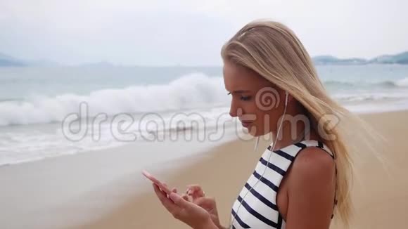 年轻漂亮的苗条女人一头金发穿着黑白连衣裙站在海岸上用智能手机视频的预览图