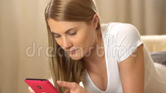 美丽迷人的年轻女人穿着白色t恤粉红色的智能手机躺在沙发上浏览互联网及视频的预览图