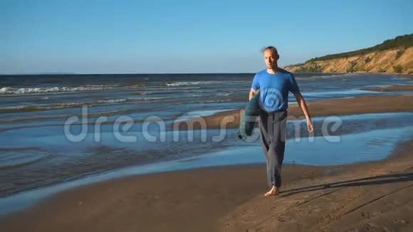 一个拿着瑜伽卷在沙滩上散步的男人视频的预览图