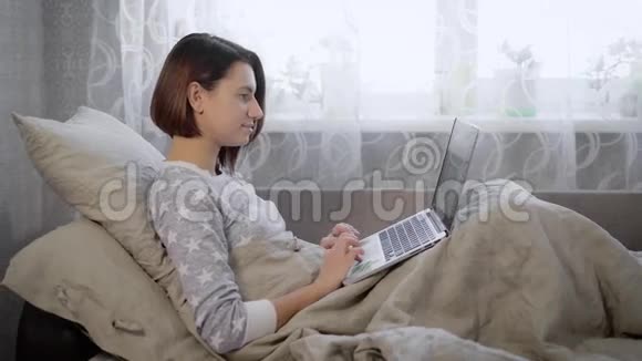 一幅美丽的黑发女人躺在床上在家里的卧室里放着笔记本电脑剪短头发的年轻女孩视频的预览图