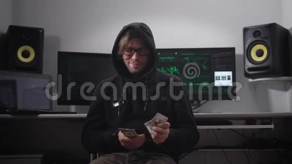 一个穿着夹克衫戴着引擎盖的人在电脑室戴眼镜手拿钱的黑客坐在椅子上一个男人视频的预览图