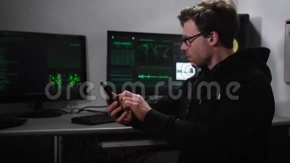 一个电脑室的黑客一个戴着太阳镜手上拿着智能手机的男人试图破解来自外国的信息视频的预览图