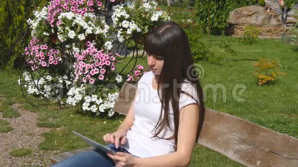 市立公园里坐在长凳上的一位年轻可爱的女士手里拿着平板电脑女孩坐在数码平板电脑上打字视频的预览图