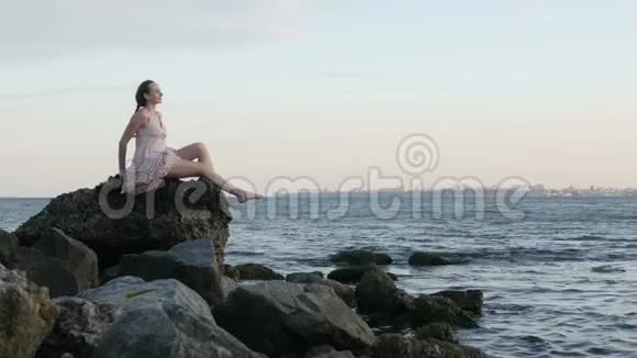 夏日时尚时髦女郎穿着漂亮的裙子坐在沙滩上的石头上视频的预览图