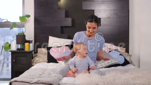 快乐的年轻妈妈微笑着和她的小儿子一起躺在家里的床上玩视频的预览图