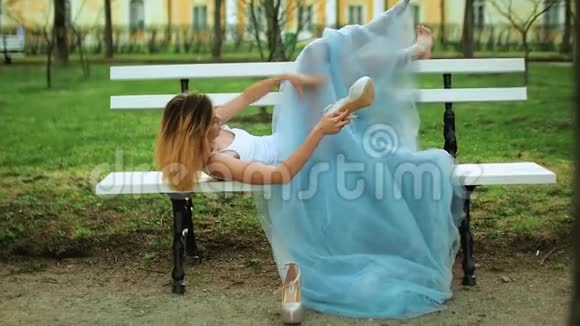 穿着白色和蓝色连衣裙的漂亮女孩光着脚躺在长凳上处理照片中摆姿势的高脚鞋视频的预览图