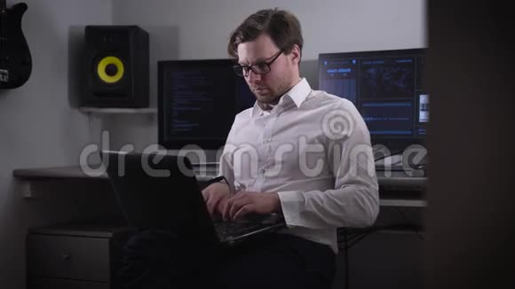 年轻的程序员穿着一件白色衬衫戴着一副时髦的眼镜在笔记本电脑上工作膝盖上有几个背景视频的预览图