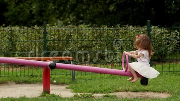 在夏日阳光明媚的日子里快乐的女孩子穿着白色裙子和衬衫在外面的跷跷板上玩耍视频的预览图