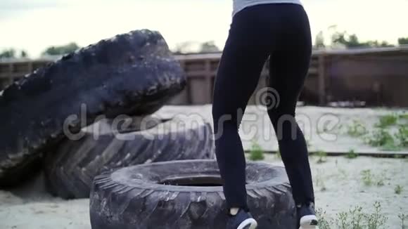 穿着灰色上衣和黑色紧身裤的运动妇女使用大型重型牵引轮进行力量练习训练视频的预览图