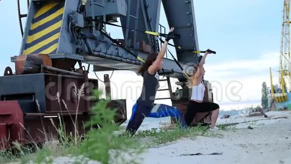 黎明时分在货物起重机附近两个年轻漂亮的运动女孩用TRX悬挂带做运动健身视频的预览图