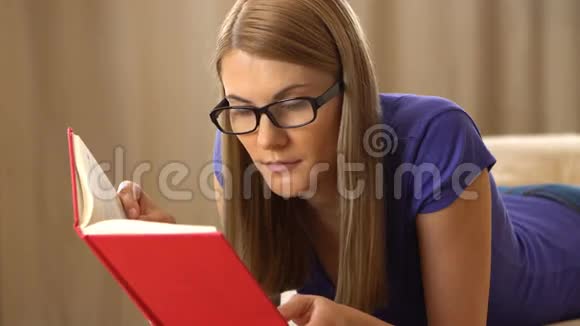 美丽迷人的年轻女人穿着紫罗兰色的t恤和一副黑色眼镜躺在沙发上看书视频的预览图