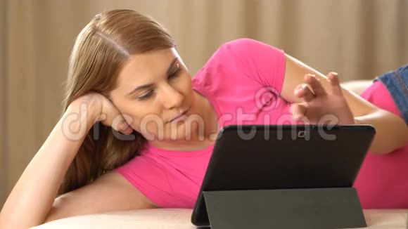 漂亮迷人的年轻女人穿着一件粉红色的t恤一台平板电脑躺在沙发上浏览互联网和微笑视频的预览图