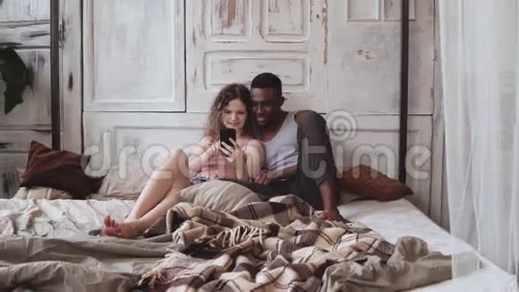 多种族夫妇穿着睡衣坐在床上使用智能手机非洲男人和欧洲女人看着照片笑着视频的预览图