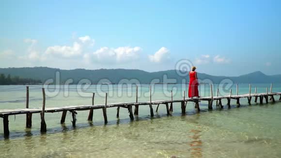 那个穿着红色连衣裙和帽子的女人走在一个长长的木制码头上慢吞吞的白色热带沙视频的预览图