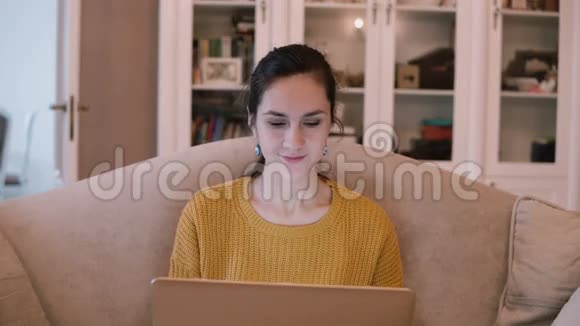 年轻女子坐在客厅里用笔记本电脑女性坐在沙发上在电脑上工作微笑视频的预览图