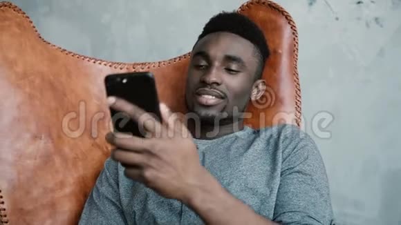 非洲年轻人坐在椅子上的肖像使用智能手机一个英俊的男人微笑着在电话里说话视频的预览图