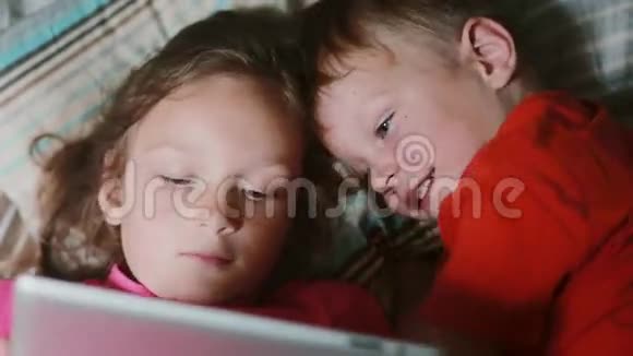 两个小孩躺在床上在触摸屏平板上看卡通兄弟姐妹一起休息视频的预览图