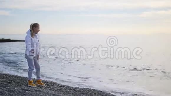 穿着夹克的年轻女子把鹅卵石抛到海里泡沫般的海浪溅上岸视频的预览图