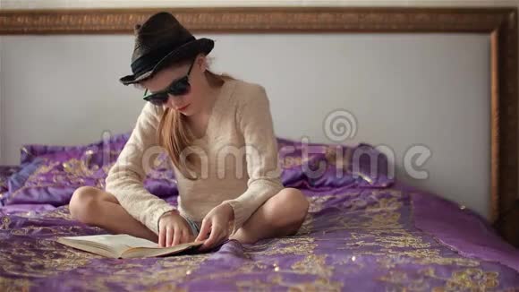 戴着墨镜和帽子的小女孩看书看着坐在床上的镜头视频的预览图