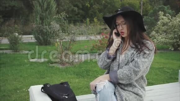 戴着黑帽子和眼镜的年轻美女在公园里打电话视频的预览图