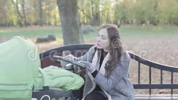 秋天一位年轻的母亲坐在公园的长凳上摇着婴儿车视频的预览图