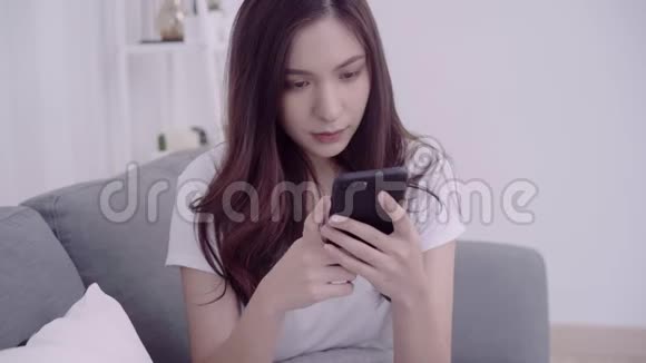 漂亮的亚洲女人在客厅里躺在沙发上用智能手机视频的预览图