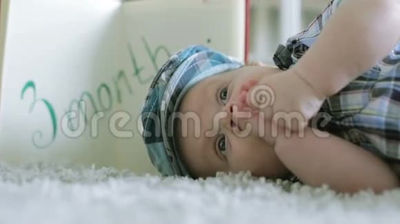 戴着方格帽的三个月大的婴儿躺在灰色地毯上吮吸着手臂视频的预览图