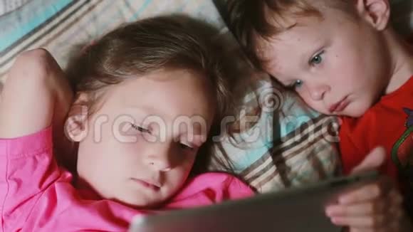 女孩和男孩躺在床上在触摸屏平板上看卡通兄弟姐妹一起休息视频的预览图