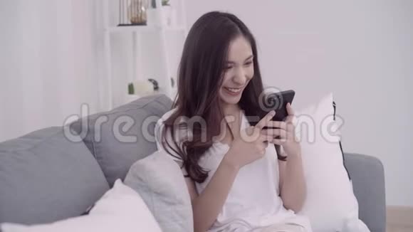 漂亮的亚洲女人在客厅里躺在沙发上用智能手机视频的预览图