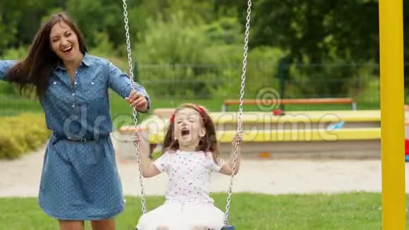 在夏日温暖的日子里微笑的美丽女人在游乐场的秋千上骑着她的女儿视频的预览图