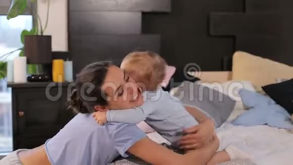 快乐的年轻妈妈微笑着和她的小儿子一起躺在家里的床上玩视频的预览图