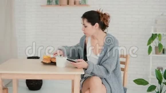 年轻女子在厨房里吃早餐用智能手机浏览互联网女孩吃羊角面包喝咖啡视频的预览图
