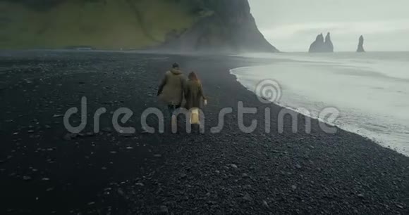 这对年轻的时髦夫妇在冰岛巨魔脚趾附近的黑色火山海滩上奔跑视频的预览图
