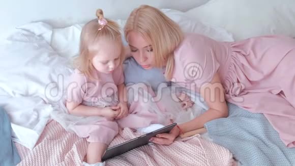 年轻迷人的金发女人用躺在床上的平板电脑教她穿着粉红色裙子的迷人小女儿视频的预览图