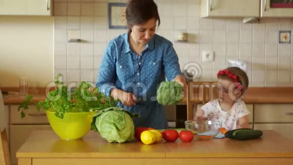 年轻的黑发女人正在厨房里切蔬菜她吃了西兰花和胡萝卜她的小女儿帮助她的母亲视频的预览图