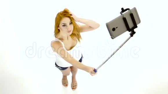 红头发的女孩用智能手机和monopod拍自拍照片视频的预览图