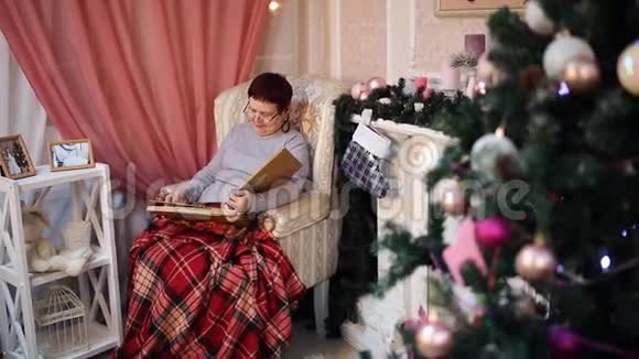 一位戴眼镜的老妇人坐在火炉旁的扶手椅上翻阅着一张带有照片的相册除夕视频的预览图